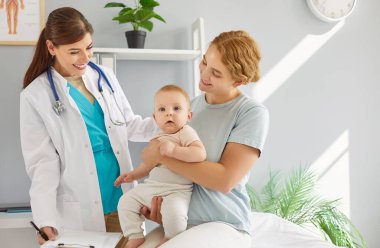 Genç, gülümseyen bir anne, parlak bir muayenehanedeki çocuk doktoruna giderken bebeğini kucağında tutuyor. Dost canlısı doktor onlarla sıcak etkileşim kurarak çocuk bakımına odaklanmış olumlu bir atmosfer yaratıyor..