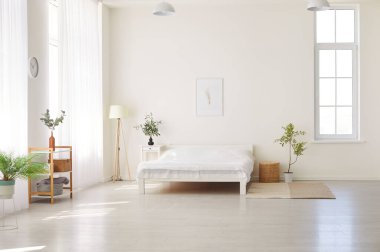 Rahat yatağı, beyaz çarşafları ve yeşil bitkileri olan ferah, güneşli bir yatak odası. Beyaz duvarları, sıcak halısı ve bir sürü penceresi olan minimalist dizaynlı modern şık yatak odası..