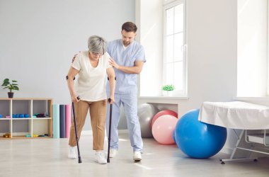 Fizyoterapist doktor, rehabilitasyon sırasında yaşlı bir hastaya koltuk değnekleriyle yürümesine yardımcı oluyor. İyileşme sürecinde hareket kabiliyetinin iyileştirilmesi ve travma veya yaralanma tedavisine odaklanılıyor..