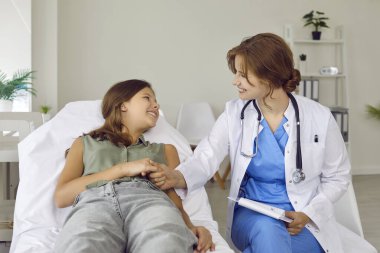 Doktor çocuğuna bakar ve ona destek olur. Dost canlısı bir çocuk doktoru ve sınav yatağında uzanan mutlu bir genç kız el ele tutuşup gülümsüyor.