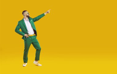 Yeşil takım elbiseli, beyaz gömlekli ve güneş gözlüklü yakışıklı şovmen sarı arka planda işaret parmağı, tam boy portre. Reklam ve pazarlama afişi.