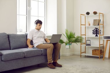 Genç adam evdeki bilgisayarını kullanıyor. Kulaklıklı bir erkek öğrenci, modern bir oturma odasında rahat bir kanepede otururken bilgisayarında bir konferans izliyor.