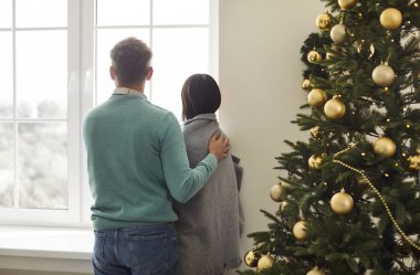 Evli bir çiftin arka plandaki görüntüsü, yeni yılı veya Noel 'i kutlarken pencereden dışarı bakarken kucaklaşmayı paylaşır. Birlikte, evde tatil sezonunun sıcaklığından ve düşkünlüğünden zevk alıyorlar..
