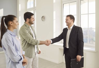 Emlakçı potansiyel ev alıcılarını el sıkışarak ve profesyonelce davranarak selamlıyor. Genç beyaz çift, danışmanlık için erkek emlakçıyla ev satın alma süreci toplantısı konusunda heyecanlı.