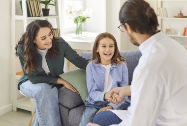 Annesiyle klinikte oturmuş gülümseyen bir çocuk tıbbi muayeneden sonra erkek doktorla el sıkışıyor. Arkadaş canlısı çocuk doktoru ve neşeli çocuk el sıkışarak başarılı tedaviyi kutluyor..