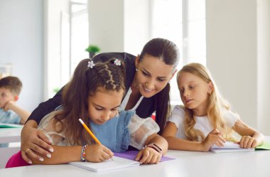 İlkokul derslerinde küçük okul kızlarına yardım eden endişeli bayan öğretmen. Gülümseyen kadın çocuklara hecelemeyi öğretiyor. Çocuklar not defterlerine yazarlar. Ortaöğretim kavramı.