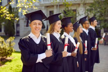 Mezuniyet günü. Mezuniyet töreni diploması olan neşeli bir üniversite öğrencisi portresi. Mezunların arka planında mezuniyet elbisesi giyen mutlu, zeki adam. Eğitim kavramı.