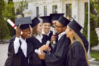 Mezuniyet elbiseleri ve kepleri içinde, üniversiteden mezun olup kampüs açıklarında konuşan, çok kültürlü, mutlu, güler yüzlü bir grup insan. Lise, üniversite, üniversite konsepti.