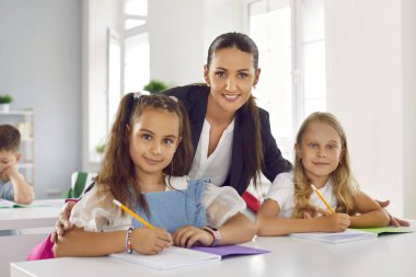 Ders sırasında sınıftaki masasında oturan iki küçük kız öğrenci ile arkadaş canlısı bayan öğretmenin portresi. Öğretmen okul ödevlerinde yardımcı olması için kızlara eğiliyor. Not defterine yazıyorlar.