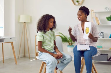 Afrikalı Amerikalı öğretmen psikologu sandalyesinde oturan öğrenci kıza gülümseyen bir ifade sergiliyor. Duyguları tanımayı öğrenmek, gençler için duygusal zekanın gelişimi..