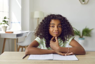 Masada elinde not defteriyle mutlu okul çocuğunun portresi. Güzel Afro-Amerikan öğrenci kız evde masada oturuyor, kameraya bakıyor ve gülümsüyor. Video çağrısı, video konferansı laptop PC ekran görüntüsü