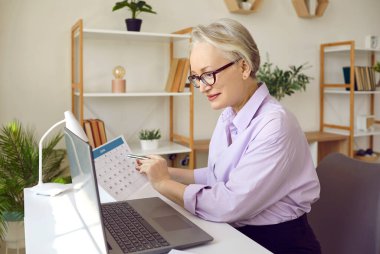 İş randevularını organize et. Seçkin üst düzey işkadını ajandasını kağıt takvim ve dizüstü bilgisayarla kontrol ediyor. İş akışı, zaman planlama ve modern teknolojilerin kullanımı kavramı. Yan görünüm.