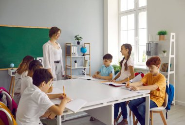 İlkokul öğrencileri ev ödevlerini uzun gün grup sınıfında birlikte yaparlar. Küçük liseli kız, büyük bir masada oturan çocuklarla çevrili sevgili bayan öğretmeninin sorularını cevaplıyor..