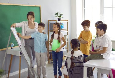 Sınıf arkadaşları okul projesinde birlikte çalışırlar. İlkokul öğrencileri ders sırasında öğretmenle birlikte beyaz tahta kullanırlar. Çocuklar sırayla tahtaya yazı yazarlar. Öğrenim ve okul kavramı.