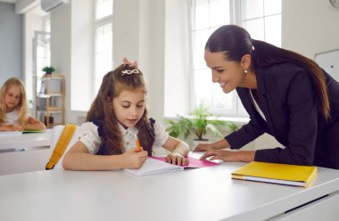 Gülümseyen arkadaş canlısı öğretmen sınıfta genç bir kıza yardım ediyor. Çalışkan ilkokul öğrencisi okul masasında oturuyor ve not defterine yazıyor, neşeli öğretmen ona yardım ediyor.