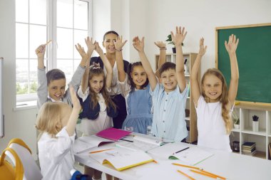 Mutlu çocuklar ve genç öğretmenler ilkokulda masa başında el ele tutuşuyorlar. Gülümsüyorlar ve kameraya bakıyorlar. Dersler arası ara, öğretmenle eğlenceli aktiviteler..
