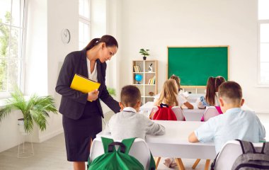 Modern beyaz bir sınıfta ilkokul öğrencileri ders veriyor. Çocuklar masalarında oturuyor ve yazı yazıyor. Genç öğretmen ders sırasında çocuklarına yardım ediyor. Okul konseptine dönelim.