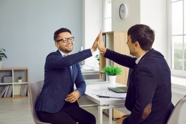 İki insan başarılı bir iş anlaşması yapar ve birbirlerine beşlik çakarlar. Takım elbiseli iki genç adam bir ofis masasında oturmuş, birbirlerine beşlik çakıyor ve gülümsüyor.