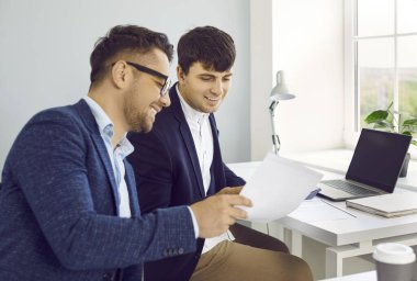 Ofiste evrak işleriyle uğraşan iki kişi. Takım elbiseli mutlu genç adamlar şirket belgelerine bakıyor, profesyonel sözleşme detaylarını inceliyor ve gülümsüyor.