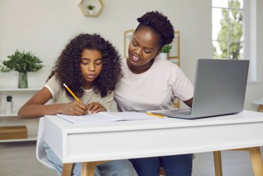 Okul ödevlerinde çocuğa yardım eden genç bir ebeveyn. Mutlu Afrika kökenli güzel anne ve küçük kızı evlerinde oturmuş, modern dizüstü bilgisayar kullanıyor ve deftere yazı yazıyorlar.