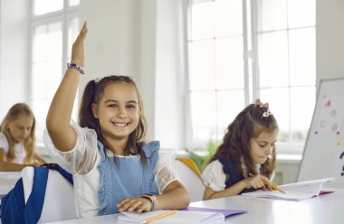 Gülümseyen zeki kız ilkokul öğrencisi öğretmen sorusuna cevap vermek için sınıf dersinde masada oturuyor. Kameraya bakıp gülümsüyor. Sınıfta eğitim süreci, ilkokul.