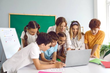 Eğitim ve teknoloji. Modern okul sınıfında kadın öğretmeni olan bir grup çocuk dizüstü bilgisayar kullanıyor. Gülümseyen öğretmen ve çocuklar modern okulda ders sırasında dizüstü bilgisayara bakıyorlar..