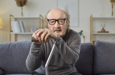 Evde bastonlu yaşlı bir adamın portresi. 60 'lı yaşlarda, gözlüklü yaşlı bir adam kanepede oturuyor, elleri bastonun üzerinde dinleniyor ve gülümsemeyen, duygusuz bir yüz ifadesiyle kameraya bakıyor.