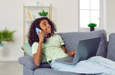 Mutlu çocuk evde koltukta oturuyor ve cep telefonuyla konuşuyor. Mutlu, rahat Afro-Amerikalı kız dizüstü bilgisayarla koltukta oturuyor, cep telefonundan arkadaşını arıyor ve gülümsüyor.