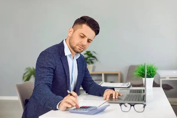 Серйозний бізнесмен в офісі обчислює щомісячні витрати, податки і підсумовує загальну суму. Людина в модному костюмі працює з ноутбуком, калькулятором і паперовими документами на робочому місці. Власна бізнес-концепція.