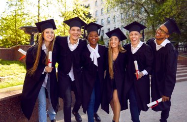 Mezuniyet gününü birlikte kutlayan çok ırklı öğrencilerin mutlu portresi. Açık havada poz veren akademik önlüklü ve havan mermili gençler. Eğitim, mezuniyet ve halk konsepti.