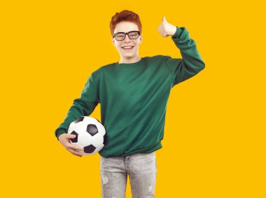 Gülümseyen, kızıl saçlı, şeffaf gözlüklü, kazak ve kot giyen çocuk başparmağını kaldırıyor ve sarı arka planda futbol topunu tutuyor. Gelecekteki futbolcunun çocukluk portresi.