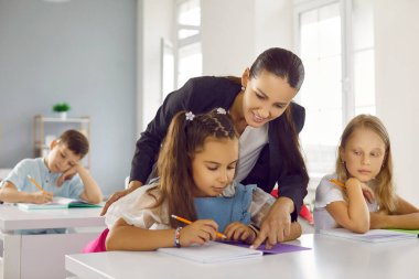 Genç, güzel esmer öğretmen, okul kızının defterindeki işi çözmesine yardım ediyor. Eğitim, ilkokul, öğrenme ve insanların konsepti. Sınıfta bir grup öğrenci yazı sınavı yapıyor..