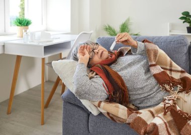 Olgun kadının sağlıkla ilgili sorunları var. Sağlıksız yaşlı bir kadın yüksek ateş ve şiddetli baş ağrısıyla evdeki kanepede yatıyor. Kadın alnını tutuyor ve termometreye bakıyor. Yaşlı sağlık konsepti.