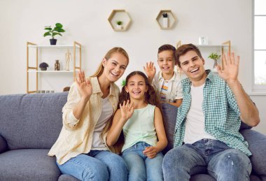 Aile görüntülü konuşma yapıyor. Evde iki çocuklu neşeli genç bir çift online video görüşmesi yoluyla akrabalarıyla iletişim kuruyor. Aile kanepeye oturur ve ellerini web kamerasının önünde sallar..