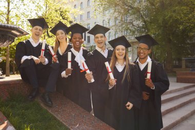 Gülümseyen bir grubun portresi mutlu çok ırklı uluslararası mezun öğrencilerin üniversite mezuniyet elbisesi içinde ve açık havada diploma tutarak sıraya dizilmeleri. Eğitim ve mezuniyet kavramı.