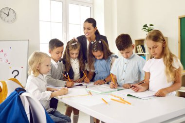 Bir grup ilkokul öğrencisi, genç bayan öğretmenle birlikte görevi tamamlıyor. Okul çocukları bir masada durur ve öğretmenlerini dinlerken not defterlerine yazarlar..
