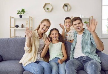 İki çocuklu genç bir aile akrabalarını karşılamak için web kamerasının önünde ellerini sallıyor. Mutlu anne, baba, oğul ve kız oturma odasında kanepede otururken görüntülü konuşma yapıyorlar..