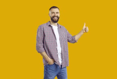 Mutlu, yakışıklı, sakallı, sıradan gömlekli, kot pantolonlu, turuncu sarı arka planda tek elini cebine atmış kameraya bakıyor, gülümsüyor ve baş parmağını kaldırıyor.