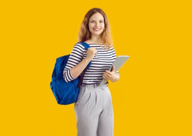 Üniversitede ya da üniversitede okuyan mutlu kız öğrencinin stüdyo fotoğrafı. Sırtında mavi sırt çantası ve sarı arka planda duran not defteriyle sıradan giyinmiş gülümseyen genç bir kadın.