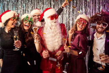 Bir grup mutlu genç arkadaş ellerinde şampanya bardaklarıyla Noel Baba kostümlü bir adamla yılbaşı partisini kutlarken eğleniyorlar. Noel tatili kavramı.