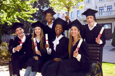 Bir grup neşeli üniversite mezunu, diplomalarıyla unutulmaz bir fotoğraf için poz veriyorlar. Akademik elbiseler ve havan tahtaları giymiş gülümseyen çok ırklı öğrenciler açık hava bankında yan yana oturuyorlar. Mezuniyet konsepti.