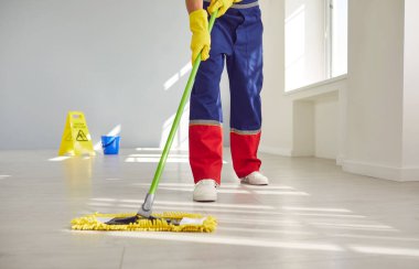 Düşük açılı, erkek hademenin bacaklarını paspas ile temizlerken çekilmiş bir fotoğrafı. Eldivenli ve üniformalı bir adam boş gri odayı ya da ofisi temizliyor. Seçici odak.