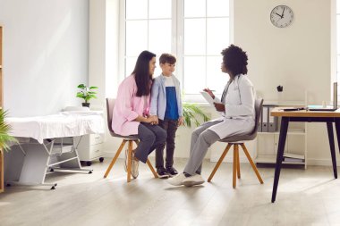 Yakışıklı çocuk ve annesi klinikteki doktoru ziyaret ediyorlar. Afro-Amerikalı kadın doktor erkek hastaya pediatrik muayene yapıyor. İlgili kadın pratisyen, çocuk doktoru hastayla konuşuyor.