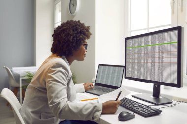 Masada oturan, bilgisayarında masaları ve çizelgeleri olan ve monitör ekranına bakan konsantre olmuş bir Afro-Amerikan kadın muhasebecinin yan görüntüsü..