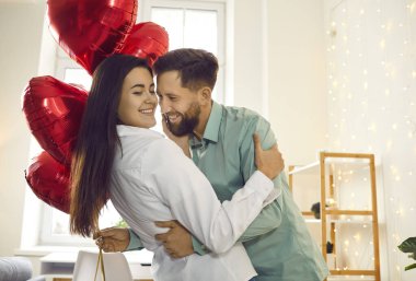 Elinde kırmızı folyo balonlarıyla gülümseyen genç, mutlu esmer adam tatildeki güzel kadını tebrik ediyor ve sarılıyor. Aşık mutlu bir çift. Dikkat, şefkat ve sevgililer günü konsepti.