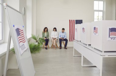 Oy verme merkezinde Amerikan bayrağıyla süslenmiş bir sıra beyaz oy kullanma kabiniyle oy kullanma merkezi. Amerikan seçmenleri sandık merkezinde oturuyorlar. Demokrasi ve seçim günü konsepti.