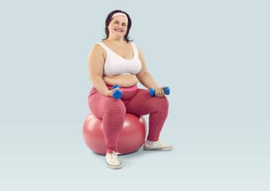 Spor kıyafetleri giyen neşeli, genç, mutlu, şişman bir kadın stüdyo mavisi arka planda münasip topun üzerinde oturan halterlerle egzersiz yapıyor. Spor, spor ve vücut pozitifliği.