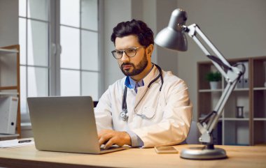 Ciddi, odaklanmış bir doktorun portresi dizüstü bilgisayarında oturup hastalarla ilgili raporları dolduruyor. Çekici doktor ofiste dizüstü bilgisayarda çalışıyor..