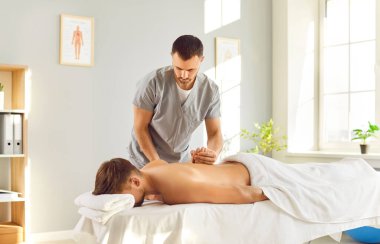 Profesyonel masör üniformalı adam klinikteki kanepede yatan rahat erkek hastaya tıbbi masaj yapıyor. Rehabilitasyon ve osteopatik tıp konsepti.