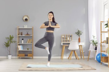 Vrksasana pozisyonunda yoga yapan spor düşkünü genç bir kadın evde spor minderi üzerinde duruyor. Mutlu rahat kız ağaçta meditasyondan zevk alıyor elleri namaste 'de, dengeyi ve dengeyi geliştiriyor..